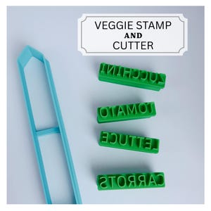 May include: A light blue elongated vegetable cutter and four green rectangular stamps with the text ZUCCHINI, TOMATO, LETTUCE, and CARROTS. A white sign at the top displays the text VEGGIE STAMP AND CUTTER in black capital letters.