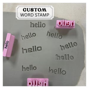 May include: A light pink custom word stamp with the text hello, shown alongside a gray clay slab featuring multiple impressions of the word hello. A white rectangular label at the top displays the black text CUSTOM WORD STAMP in a serif font.