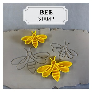 May include: The image displays bee-shaped stamps in yellow and gray. The text "BEE STAMP" is in a white rectangular frame. The stamps are on a light brown surface, with two yellow bee stamps and two gray outline stamps.