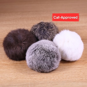 May include: Four fluffy cat toys in shades of brown, gray, and white are arranged on a wooden surface. The toys are round and made of soft, textured material. A red rectangle with the words "Cat-Approved" is in the upper right corner.