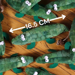 Peut inclure: Un groupe de jouets en peluche d'alligator vert avec des yeux blancs. Les alligators sont dispos&eacute;s sur une surface en bois. Une r&egrave;gle indique que la longueur d'un des alligators est de 16,5 cm.