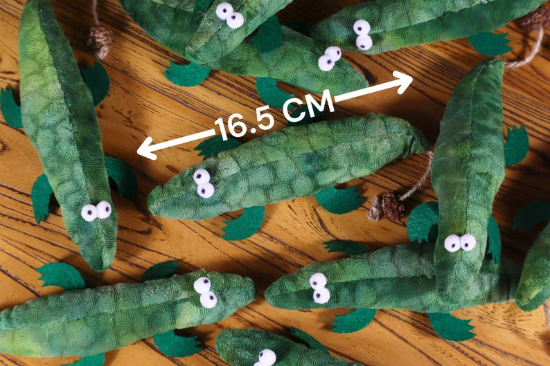 Peut inclure: Un groupe de jouets en peluche d'alligator vert avec des yeux blancs. Les alligators sont dispos&eacute;s sur une surface en bois. Une r&egrave;gle indique que la longueur d'un des alligators est de 16,5 cm.