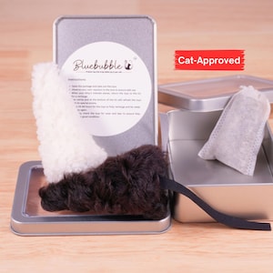 Op de afbeelding: Een zilverkleurig metalen blik met een wit en een donkerbruin pluche speeltje, met een klein wit stoffen zakje in een apart open blik. Het deksel bevat de tekst Bluebubble en instructies, met een rood label met de tekst Cat-Approved.