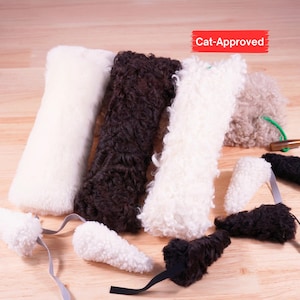 Peut inclure: Une collection de jouets en peluche à texture bouclée en blanc, brun foncé et beige. Les articles comprennent des formes rectangulaires longues et des pièces triangulaires plus petites avec des rubans. Une étiquette rouge porte le texte Cat-Approved.