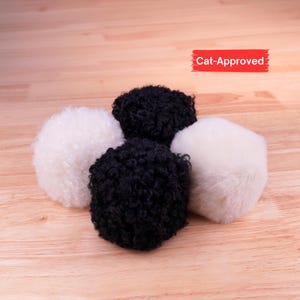 May include: Four spherical pet toys arranged in a cluster, featuring two white fuzzy balls and two black curly-textured balls. A red rectangular label in the upper right corner displays the white text Cat-Approved in a bold, sans-serif font.