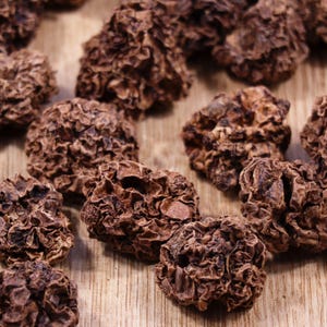 May include: Close-up of a pile of dried, dark brown, wrinkled, and textured fruit. The fruit is arranged on a light brown wooden surface.