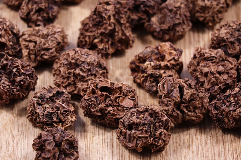 May include: Close-up of a pile of dried, dark brown, wrinkled, and textured fruit. The fruit is arranged on a light brown wooden surface.