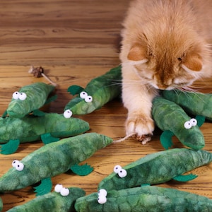 Peut inclure: Un chat roux joue avec un ensemble de jouets en peluche en forme d'alligator vert. Les jouets en forme d'alligator ont des yeux globuleux et sont faits d'un tissu doux et duveteux.