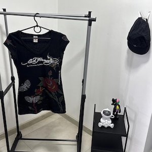 May include: Black V-neck t-shirt with a floral and butterfly design, hanging on a clothing rack. The shirt features the text "Ed Hardy by Christian Audigier". A black cap hangs on the wall. A small black shelf holds two figurines.