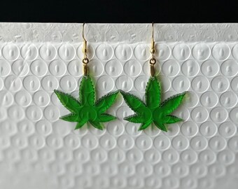 May include: A pair of green marijuana leaf earrings with gold-colored hooks. The earrings are made of a translucent green material and have a detailed leaf design. The background is a white, textured surface.