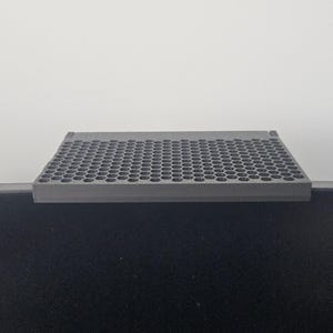 May include: A gray, rectangular, perforated shelf, likely for a monitor or desk. The shelf is made of a solid material and has a grid of small, evenly spaced holes. It sits atop a black surface, with a white background.
