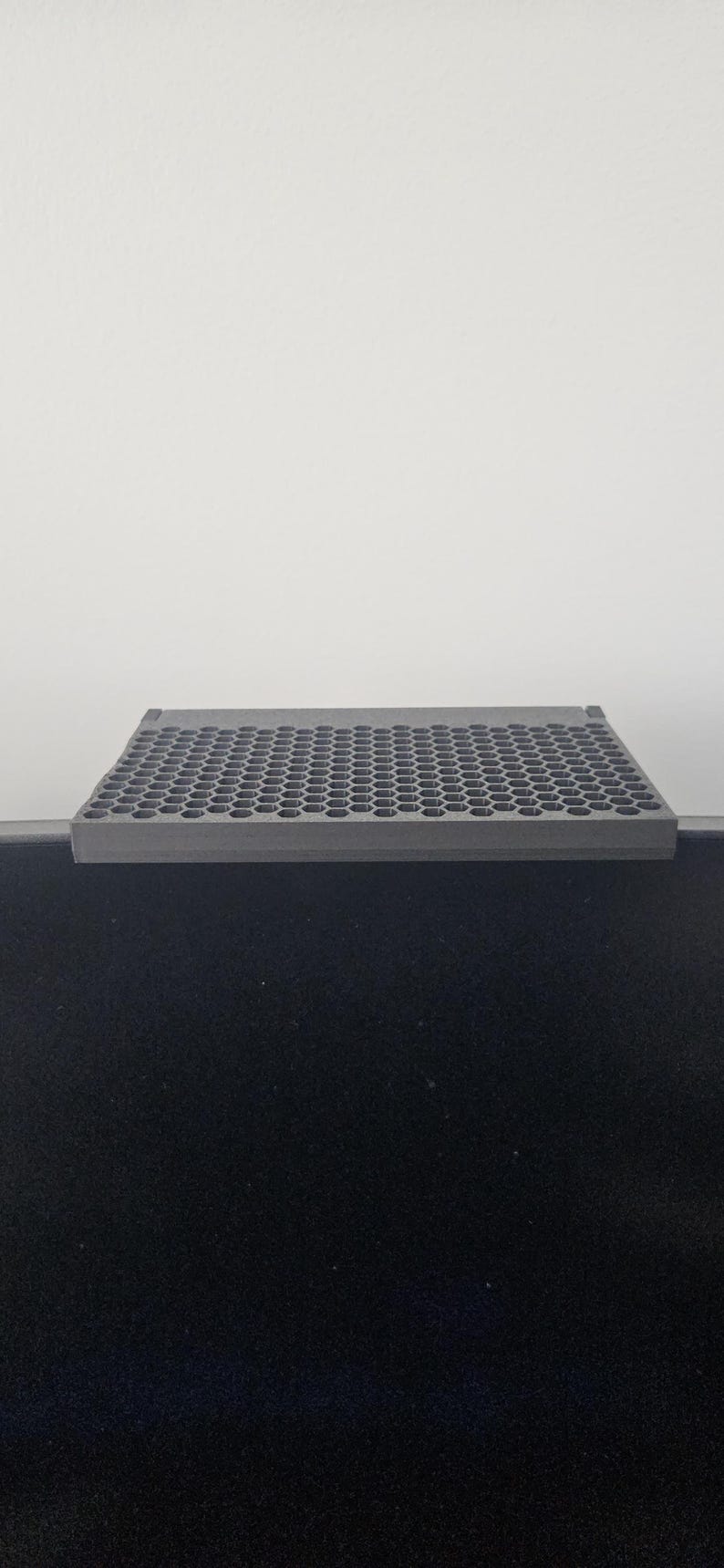 May include: A gray, rectangular, perforated shelf, likely for a monitor or desk. The shelf is made of a solid material and has a grid of small, evenly spaced holes. It sits atop a black surface, with a white background.