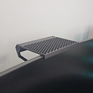 Könnte beinhalten: Ein schwarzes Monitorregal mit perforierter Oberfläche, zur Befestigung an der Oberseite eines Computermonitors. Das Regal besteht aus einem dunkelgrauen Material und hat ein schlichtes, funktionales Design.