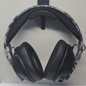 May include: A black and gray camouflage patterned headset with black earcups hanging on a black wall mount.