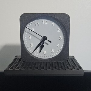 May include: A square, gray analog clock with a white face and black hands. The clock sits on a gray, textured platform. The background is a plain white wall.