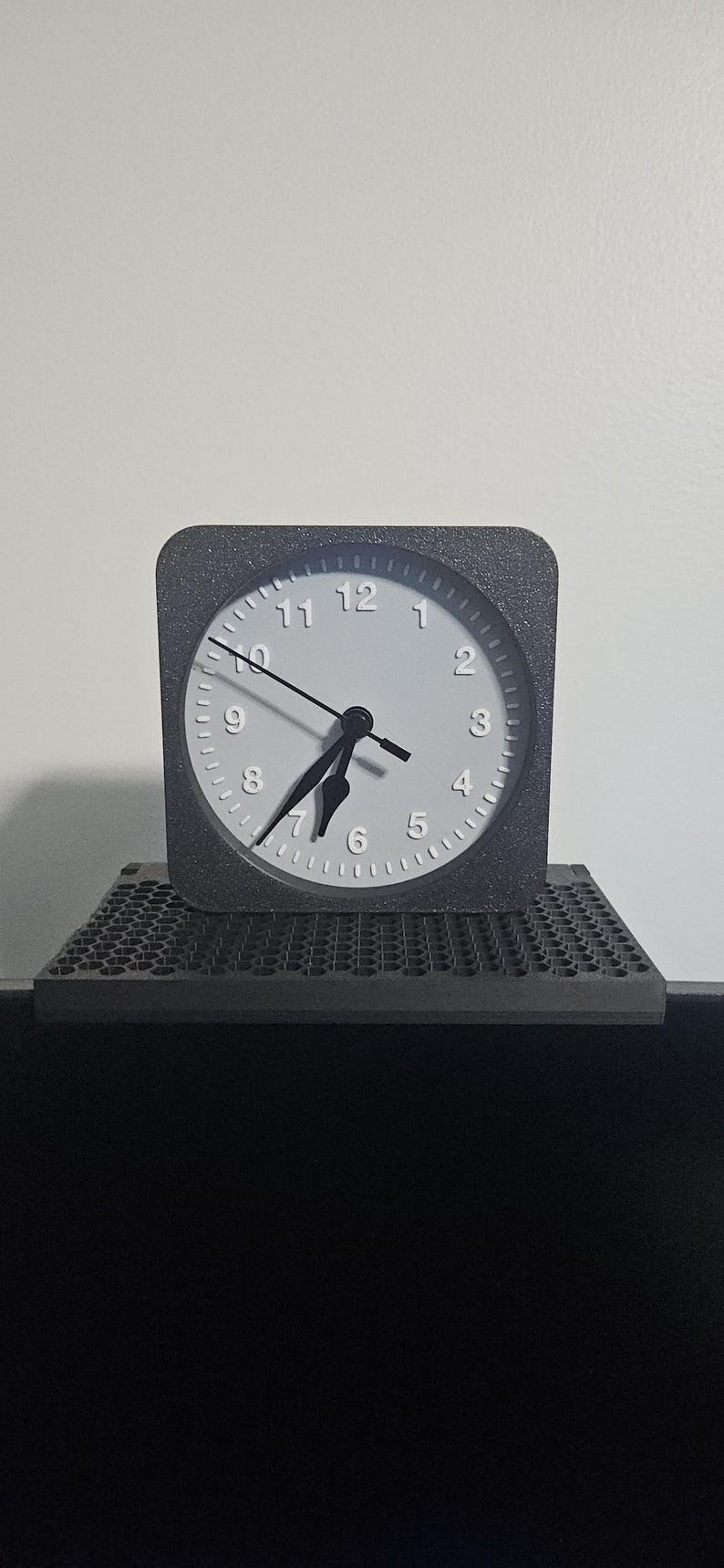 May include: A square, gray analog clock with a white face and black hands. The clock sits on a gray, textured platform. The background is a plain white wall.