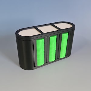 May include: A black rectangular container with three sections, each filled with stacks of bright green circular objects. The top of the container has three white rectangular openings. The design is modern and minimalist.