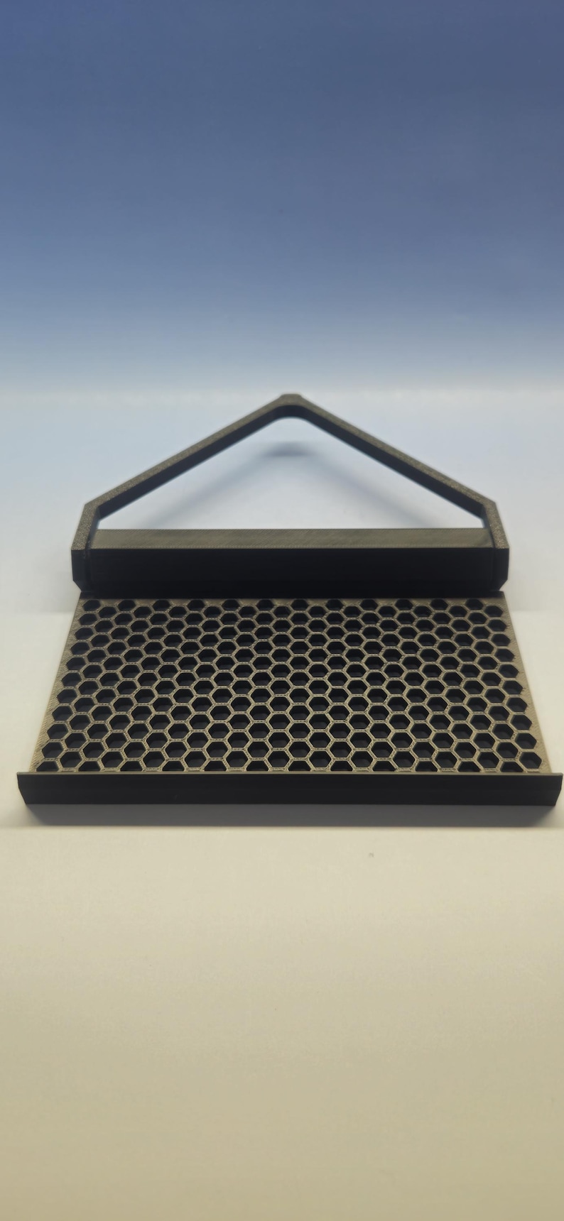 May include: A black and gray honeycomb-patterned tray with a handle. The tray has a rectangular base with a series of hexagonal holes. The handle is a triangular shape. The product is likely a kitchen or home decor item.