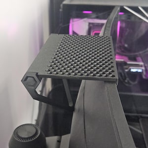 May include: A black computer monitor with a textured, square-patterned top. The monitor has a sleek, modern design with a hexagonal support structure. The background shows a glimpse of a computer's internal components.