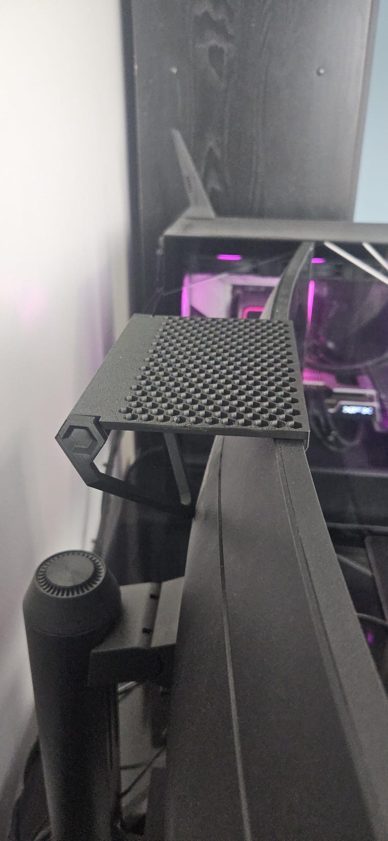 May include: A black computer monitor with a textured, square-patterned top. The monitor has a sleek, modern design with a hexagonal support structure. The background shows a glimpse of a computer's internal components.