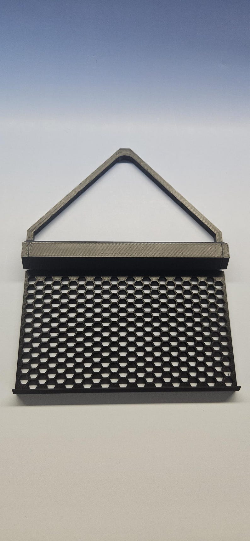 May include: A dark gray, 3D-printed object with a honeycomb pattern and a triangular handle. The object has a rectangular base and a series of hexagonal openings. The handle is a triangle shape.