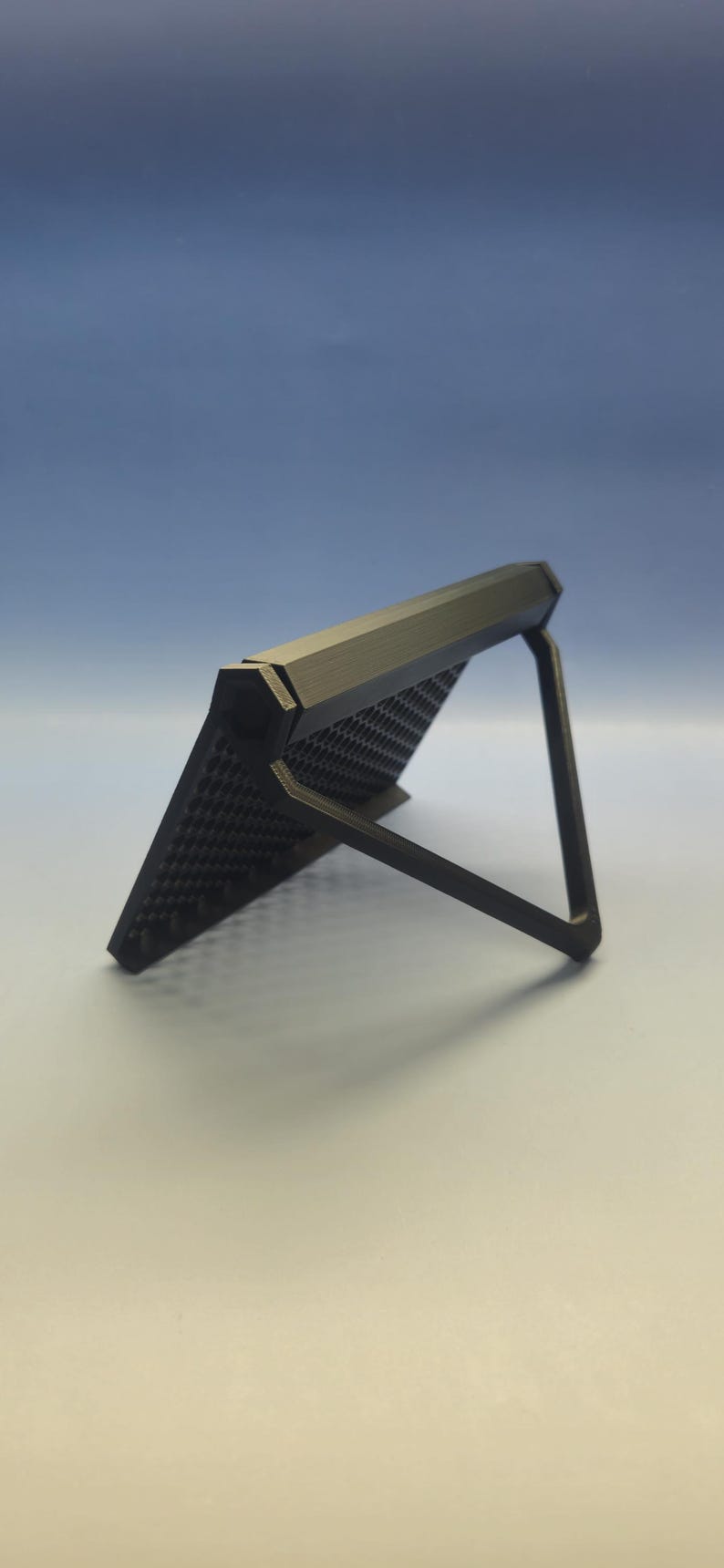 May include: A black, angled phone stand with a textured surface. The stand is supported by a thin, angled leg. The background is a gradient of blue and white.