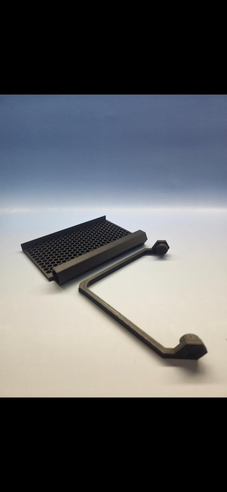 May include: A dark gray, 3D-printed component with a honeycomb-patterned grill and an L-shaped support arm. The arm has hexagonal end pieces. The items are set against a white surface with a blue gradient background.