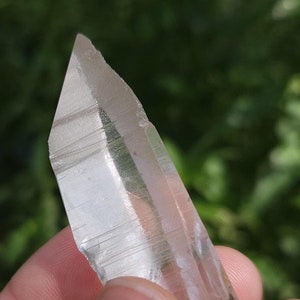 Op de afbeelding: Een heldere kwartskristalpunt met strepen, vastgehouden in een hand tegen een groene achtergrond.