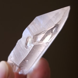 May include: A clear quartz crystal with a pointed tip and a flat base. The crystal is held in a hand.