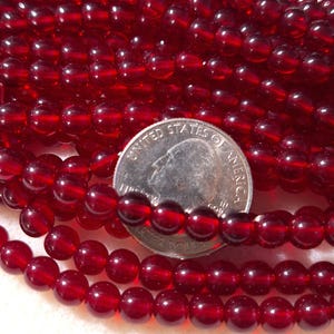 May include: A collection of deep red, translucent, spherical beads arranged in multiple rows. A United States quarter coin is placed among the beads to provide a sense of scale, showing the relative size of the small, polished, circular items.