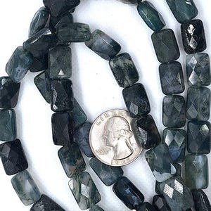 May include: A strand of faceted rectangular moss agate beads in shades of dark green and black. A silver-colored United States quarter coin is placed among the beads to provide a sense of scale for the individual stone dimensions.