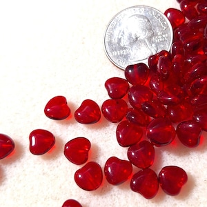 May include: A collection of translucent red heart-shaped beads scattered on a white surface. A U.S. quarter coin is visible, providing a size reference. The beads are glossy and appear to be made of glass or a similar material. The image is well-lit, highlighting the beads' color and shape.