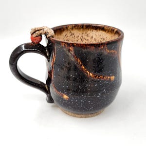 Puede incluir: Una taza de cerámica hecha a mano con un exterior marrón oscuro y un interior marrón claro moteado. La taza presenta un asa única con un pequeño acento redondo rojo. La taza está diseñada para bebidas calientes.