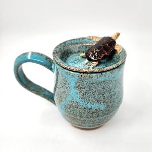 Pode incluir: Uma caneca de cerâmica turquesa com tampa a condizer, com uma figura de tartaruga castanha no topo. A caneca tem uma textura salpicada e uma pega curva. A caneca tem cerca de 10 cm de altura.