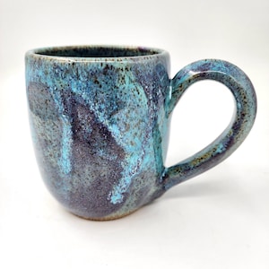 Può includere: Una tazza in ceramica fatta a mano con una smaltatura marmorizzata unica nei toni del turchese, blu e viola scuro. La tazza ha un corpo arrotondato, un manico robusto e una superficie leggermente strutturata.