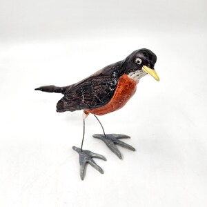 May include: A handcrafted bird figurine with a black head and back, a reddish-orange breast, and a yellow beak. The bird stands on wire legs with gray feet, set against a white background. This decorative piece is a charming addition to any home decor.