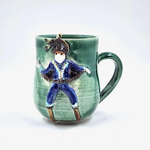 Pode incluir: Uma caneca de cerâmica verde-azulado com uma figura esculpida de uma pessoa em um traje azul e preto. A figura tem um chapéu com penas e está posando com os braços estendidos. A caneca tem uma alça curva e um acabamento brilhante.