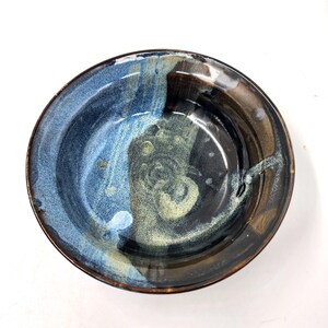 Op de afbeelding: Een handgemaakte keramische kom met een uniek glazuur. De kom heeft een mix van blauwe, bruine en crèmekleuren, waardoor een marmer effect ontstaat. De rand heeft een donkerdere bruine tint. Deze decoratieve kom is geschikt om te serveren of tentoon te stellen.