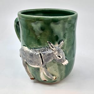 May include: A ceramic mug with a glossy, mottled green glaze featuring a three-dimensional, sculpted grey donkey attached to the exterior. The donkey is depicted in profile, facing right, with a white muzzle and a textured mane.