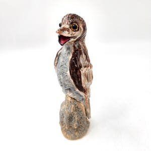 Può includere: Statuetta di uccello in ceramica con un design dettagliato e strutturato. L'uccello ha testa e ali marroni, corpo grigio e bocca rossa. È appollaiato su una base simile a una pietra. La statuetta è un oggetto decorativo.