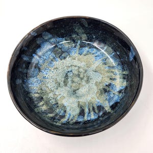 Puede incluir: Un cuenco de cerámica hecho a mano con un exterior azul oscuro y un interior moteado con tonos de azul, crema y gris. El cuenco tiene un acabado brillante y una forma ligeramente irregular, mostrando su calidad artesanal.