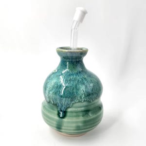May include: A handcrafted ceramic water pipe with a teal and green glaze. The pipe has a bulbous body and a slender neck, with a clear glass stem and a white connector.