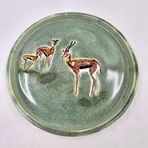 Può includere: Un piatto rotondo in ceramica con una smaltatura verde salvia maculata. Il centro presenta una scena dipinta a mano di due gazzelle marroni, una in piedi e una più piccola accanto, rese in tonalità di marrone, bianco e nero con pennellate visibili.