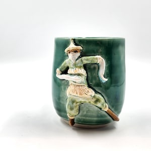 Op de afbeelding: Een teal keramische mok met een verhoogd reliëf van een figuur in een groene outfit en een hoed. De figuur staat in een dynamische pose, met uitgestrekte armen. De mok heeft een afgeronde vorm en een gladde afwerking.