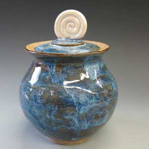 Puede incluir: Un tarro de cerámica hecho a mano con tapa. El tarro es de color azul oscuro con detalles marrones y beige. La tapa tiene un borde beige y un asa en espiral blanca. El tarro probablemente se utiliza para almacenamiento o decoración.