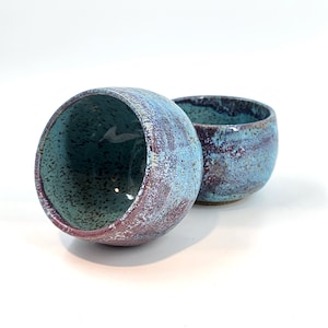 Pode incluir: Duas tigelas de cerâmica com um esmalte azul e roxo salpicado. As tigelas são redondas e têm uma ligeira borda.