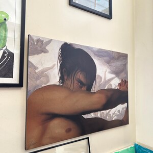 May include: A canvas print of a man with dark hair and a bare chest, looking down with a serious expression. He is surrounded by a cloudy background with a faint image of people in the distance.