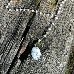 May include: A white and gray agate pendant hangs from a silver chain with white beads. The chain is draped over a piece of weathered wood.