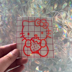 May include: Red vinyl sticker of Hello Kitty eating a strawberry. The sticker is on a white sheet of transfer paper with a grid pattern.