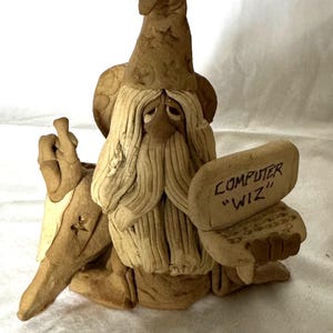May include: A tan clay figurine of a wizard wearing a pointed hat with star cutouts, a long beard, and a mustache. The wizard holds a small laptop with the text "COMPUTER WIZ" written on the screen and has one hand raised with fingers crossed.